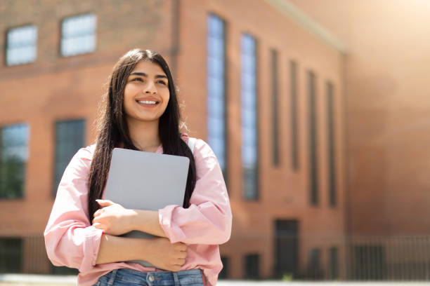 Cheerful young girl student holding a closed laptop, looking upwards thoughtfully beside a university building