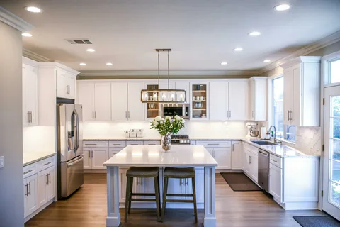 Spacious modern kitchen with white cabinets, a large central island with two stools, stainless steel appliances, pendant lighting above the island, and large windows allowing natural light to fill the room.