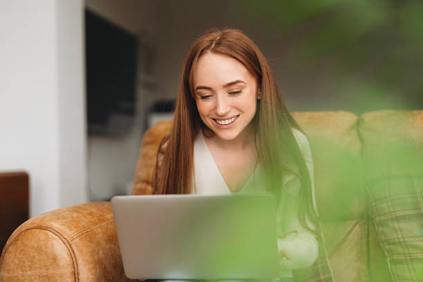 Smiling young woman engages with her laptop while sitting on a leather couch, surrounded by a warm and inviting indoor atmosphere during daylight.