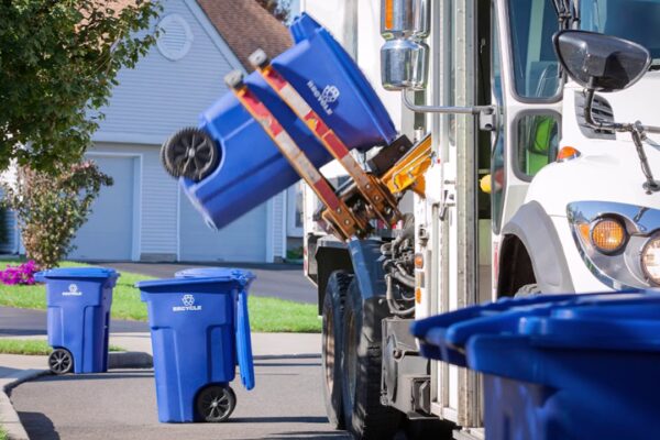 Recycling truck empties blue bins with a mechanical arm on a suburban street.