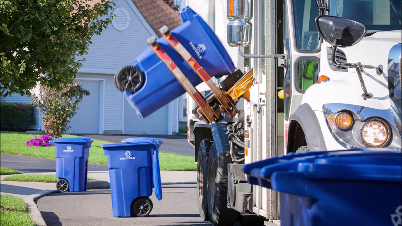 Recycling truck empties blue bins with a mechanical arm on a suburban street.